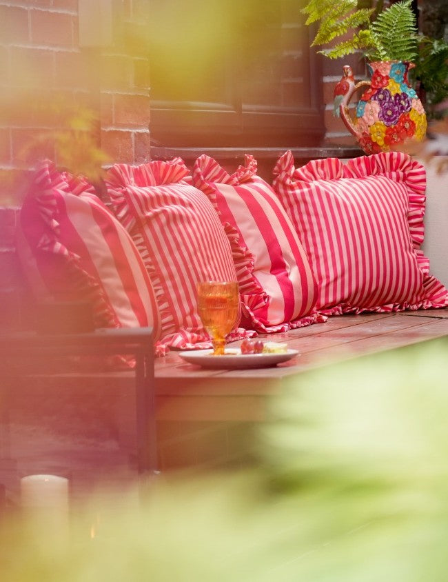 Bright cherry and peach striped ruffled cocktail cushions on a wooden outdoor bench, with an ochre glass and plate in the foreground and a brick wall behind.
