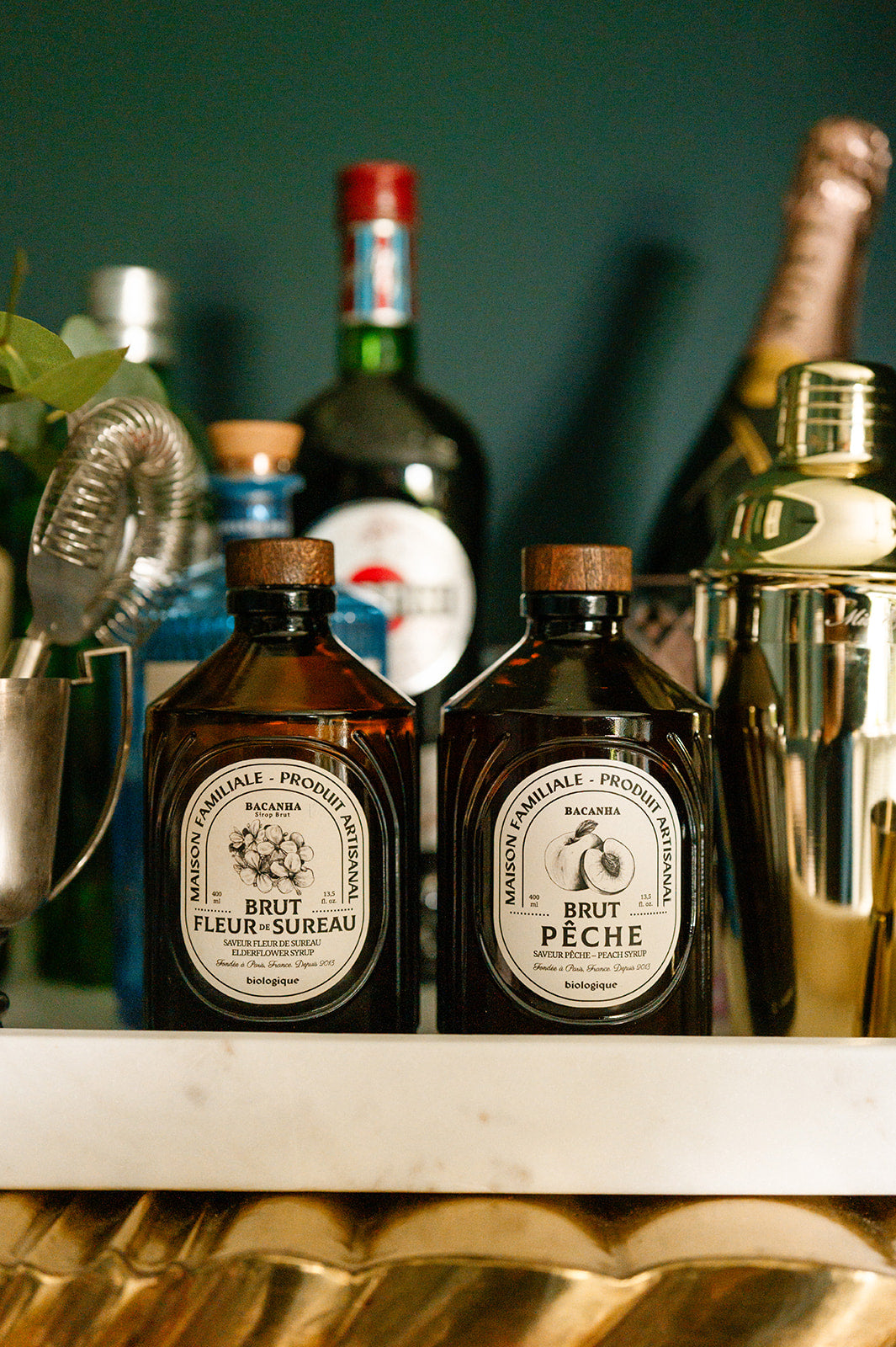 Two bottles of Fleur d'Etreau and Brut Peche on a gold art deco sideboard with cocktail shakers and bottles in the background.