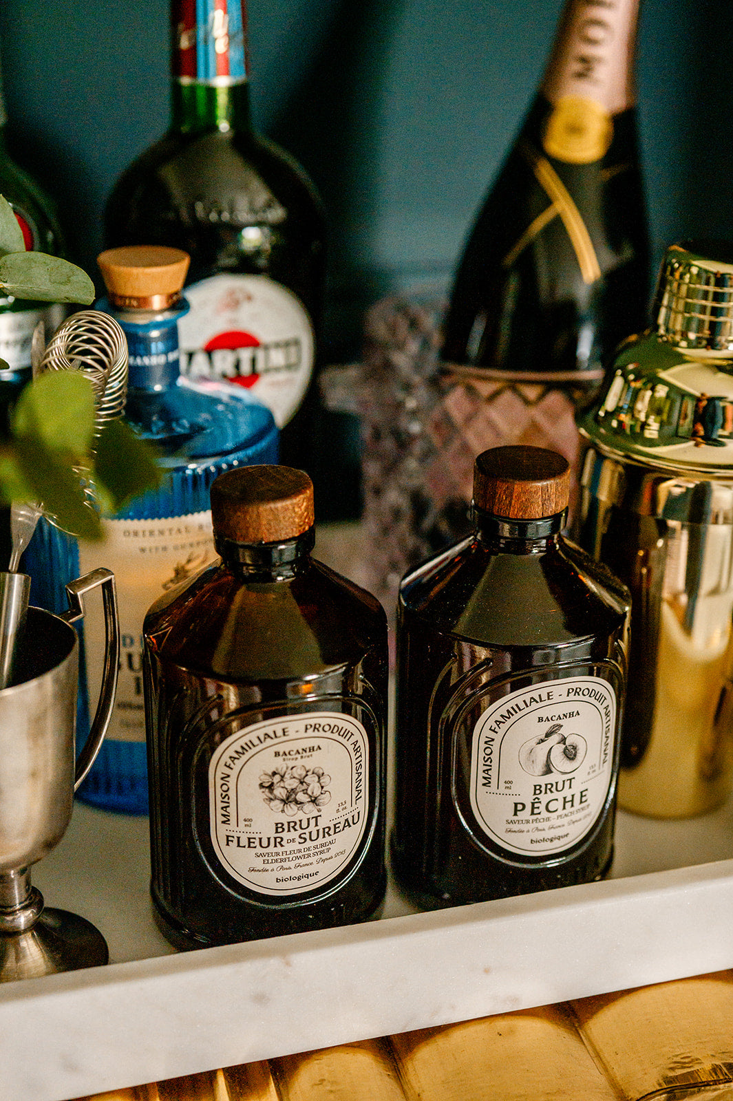 Two bottles of Fleur d'Etreau and Brut Peche on a gold art deco sideboard with cocktail shakers and bottles in the background.