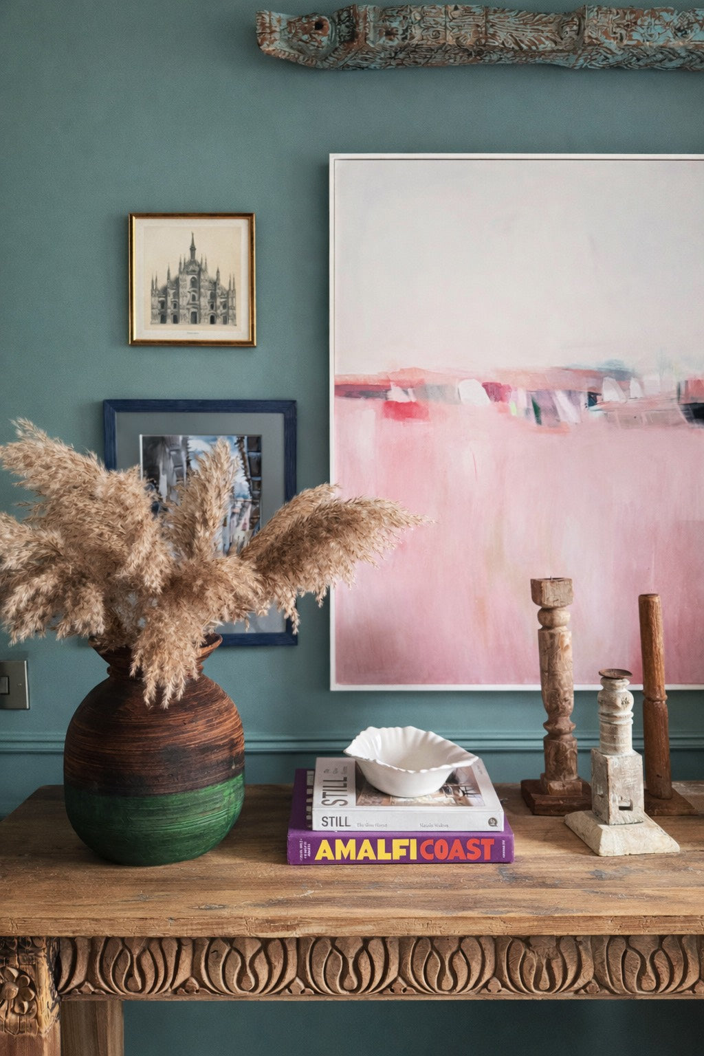 Decorative setup with a vase of pampas grass, books, and reclaimed wood candlesticks in front of a colourful painting on a vintage Indian wooden table against a teal wall.