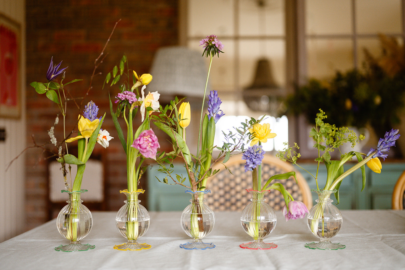 Five small glass vases with flowers on a table in a home setting