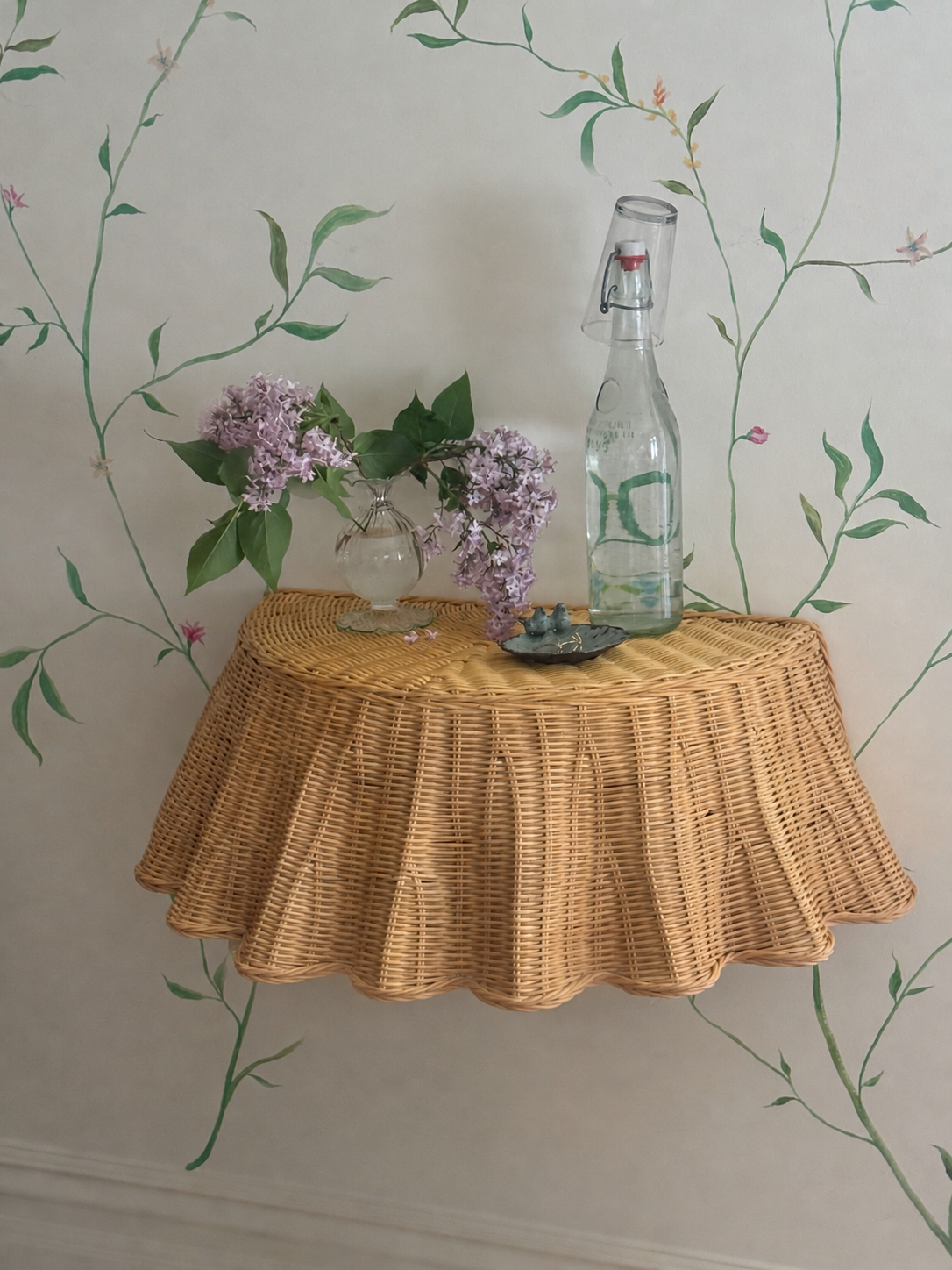 Floating rattan skirted table with fresh lilac, a glass bottle with water and a glass, verdigris trinket dish against  hand painted botanical wall mural