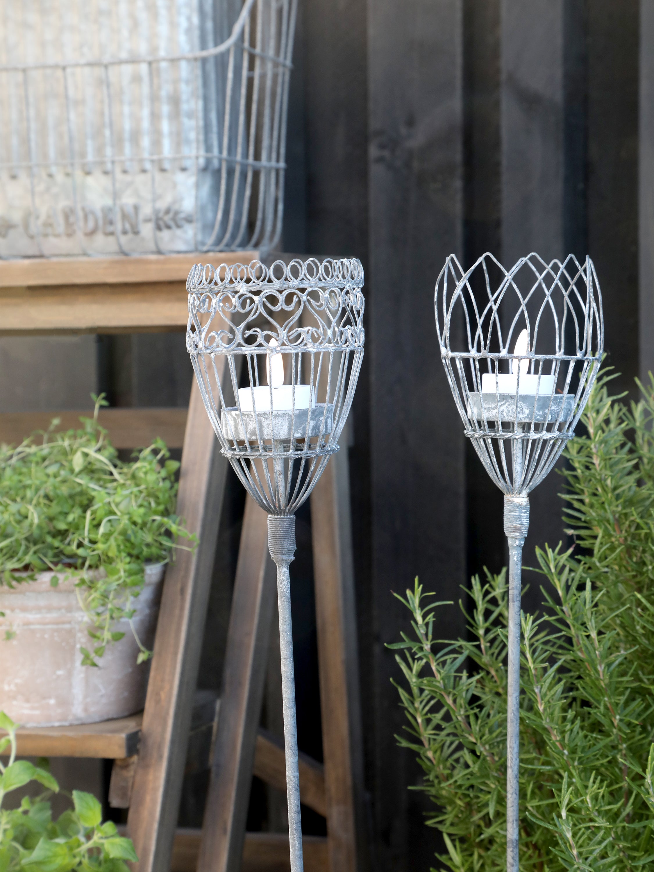 2 antique grey metal garden candle spears in front of dark wood wall and green plants