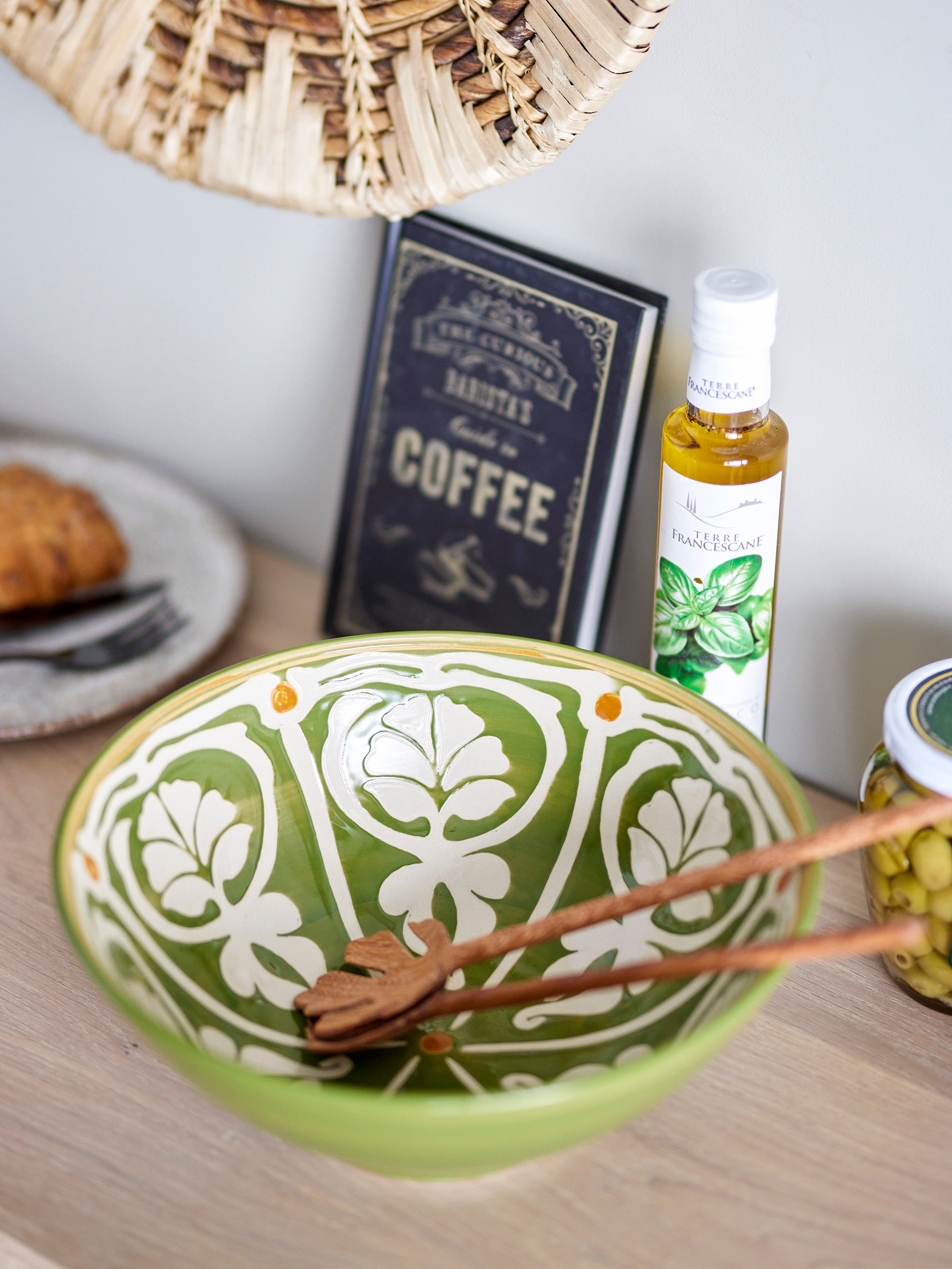 Close up of handpainted olive and cream bowl with ochre touches with wooden utensils on wood surface with condiments