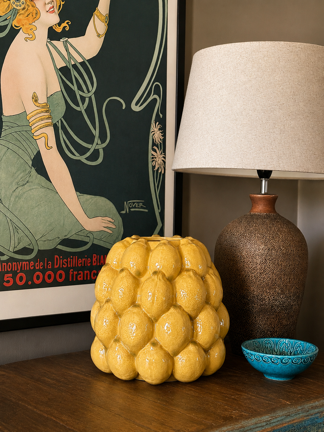 Decorative yellow lemon glazed vase, brown textured lamp, and blue bowl on a wooden surface with a framed artwork in the background.