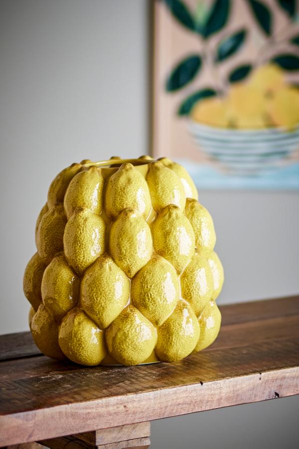 Yellow ceramic vase shaped like a stack of lemons on a wooden surface with a painting in the background.