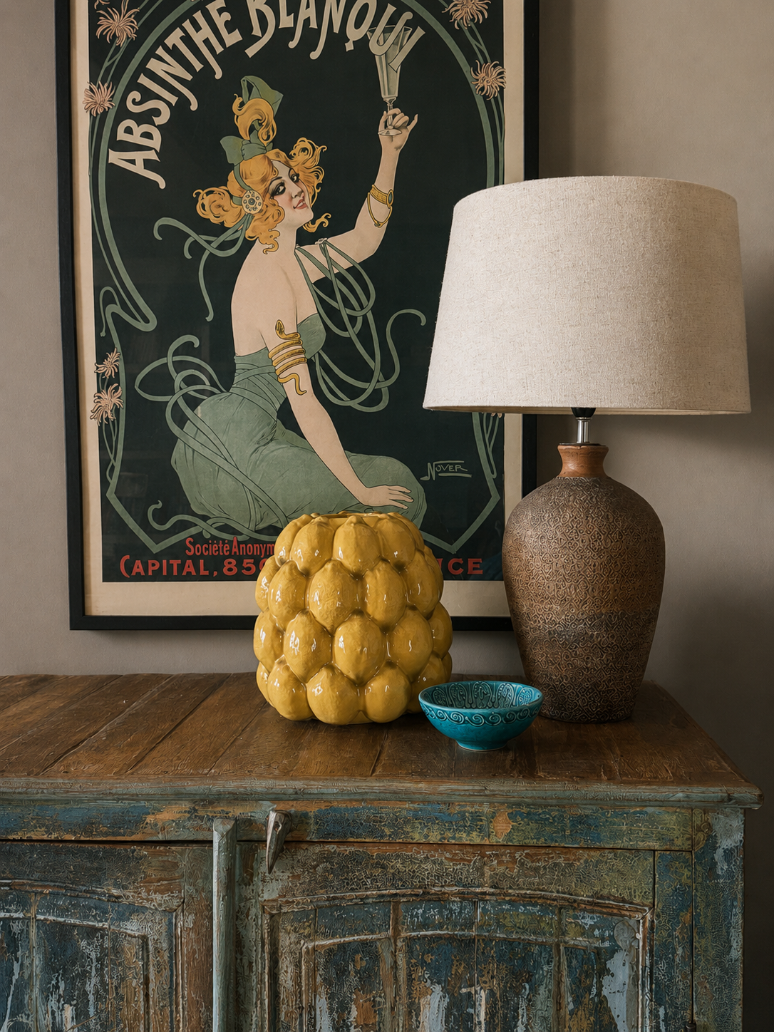 A decorative vignette with a lemon ceramic vase, textured brown lamp base with linen shade and small turquoise blue bowl on a vintage Indian turquoise cabinet with a vintage French advertising poster in the background