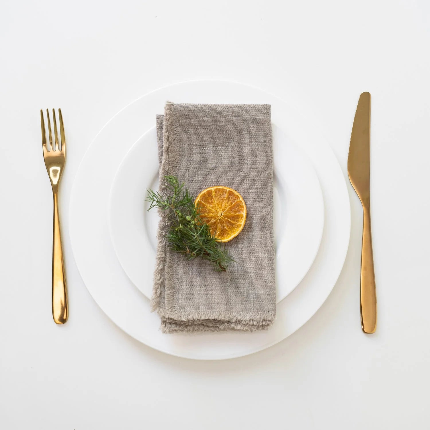 Overhead view of a place setting with a white dinner plate and gold cutlery with natural linen napkin with fringes folded with a sprig of rosemary and slice of dried orange