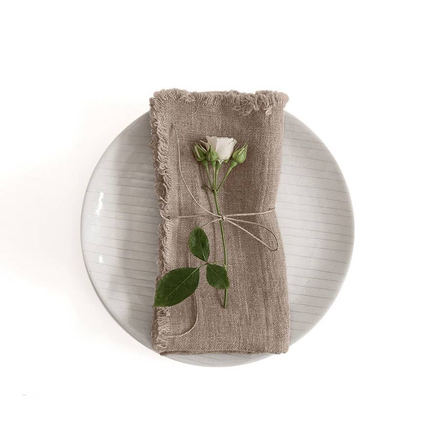 This image displays a set of two elegant linen napkins with fringes, presented in a natural beige color. The napkins are beautifully soft and are artfully arranged on a plate, showcasing their luxurious texture.