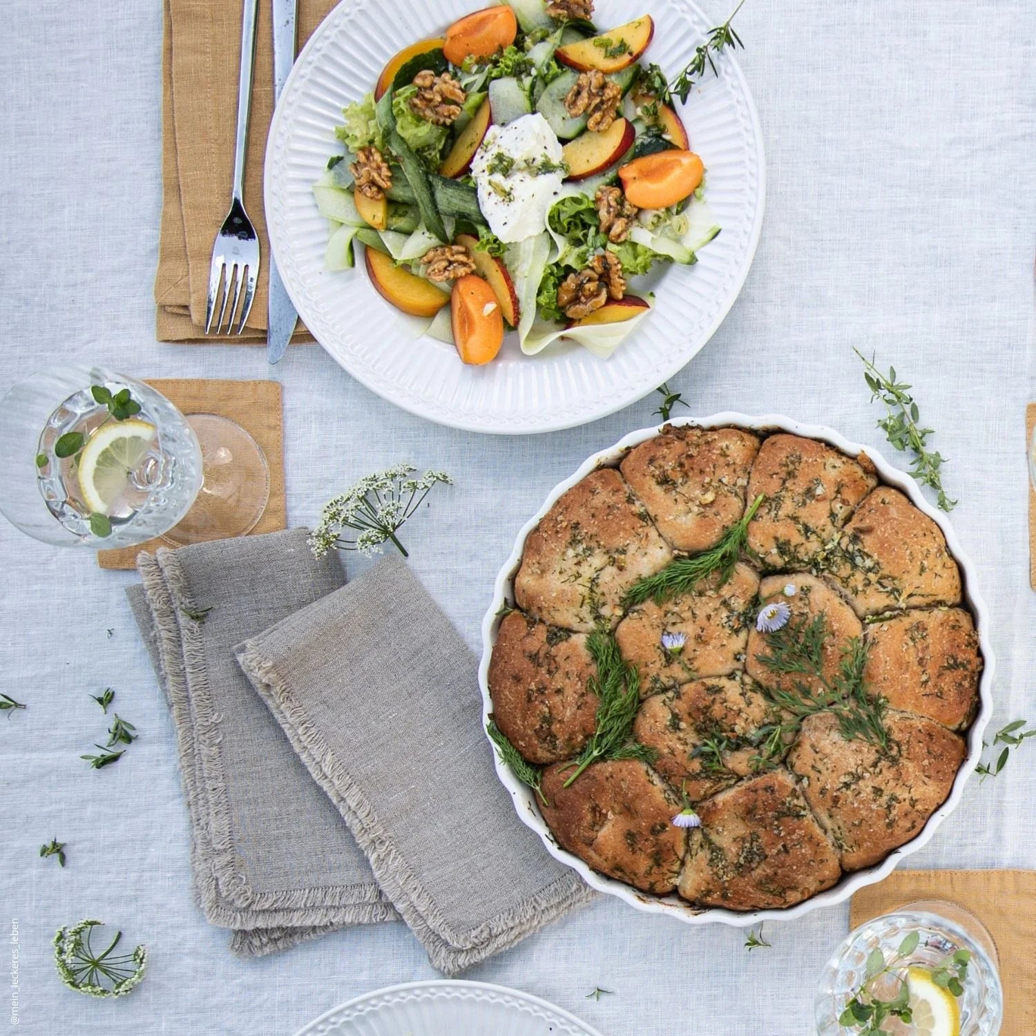 Overhead view of a table of food arranged on a light coloured linen tablecloth with natural linen napkins with fringes next to a platter 