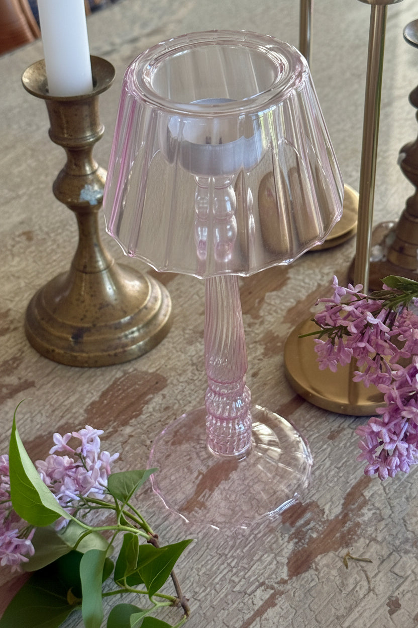 Pink glass t light stand with fluted stem, pleated shade and frilled round base on an old white textured wood table next to fresh lavender and old brass candlesticks