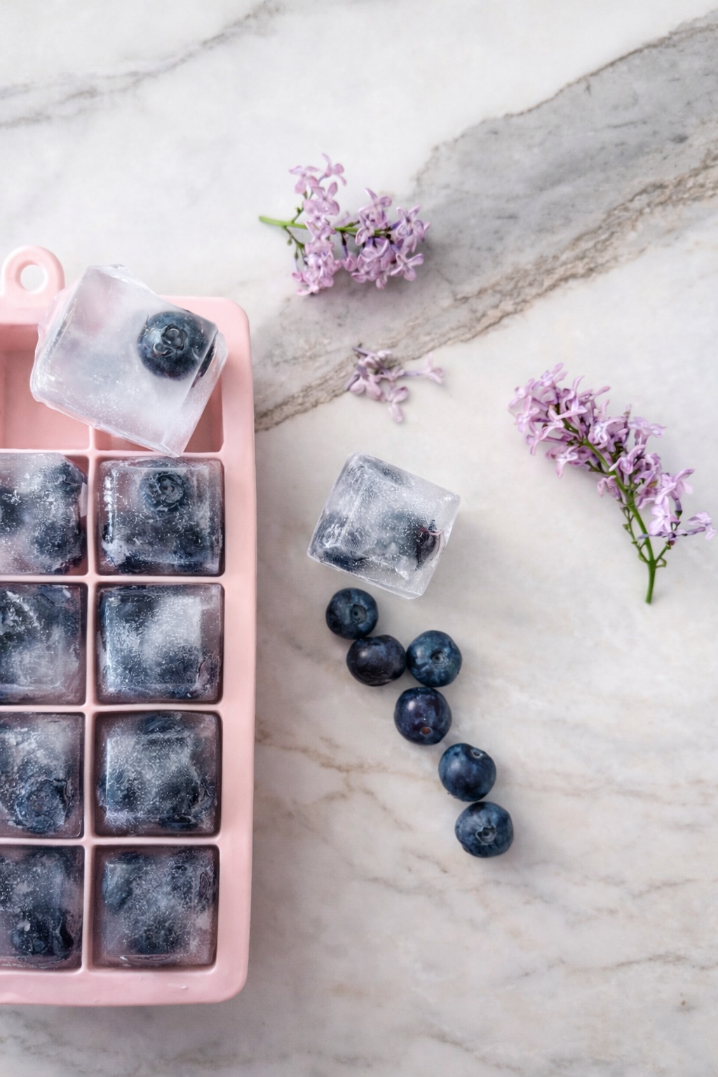 Pink ice cube tray with blueberries and purple flowers on a marble surface