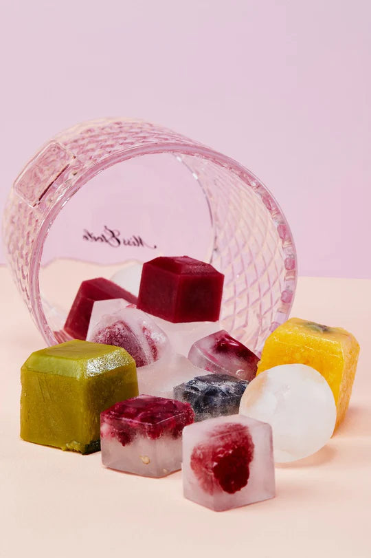 Rose ice bucket on its side with square ice cubes spilling out of it on pink surface