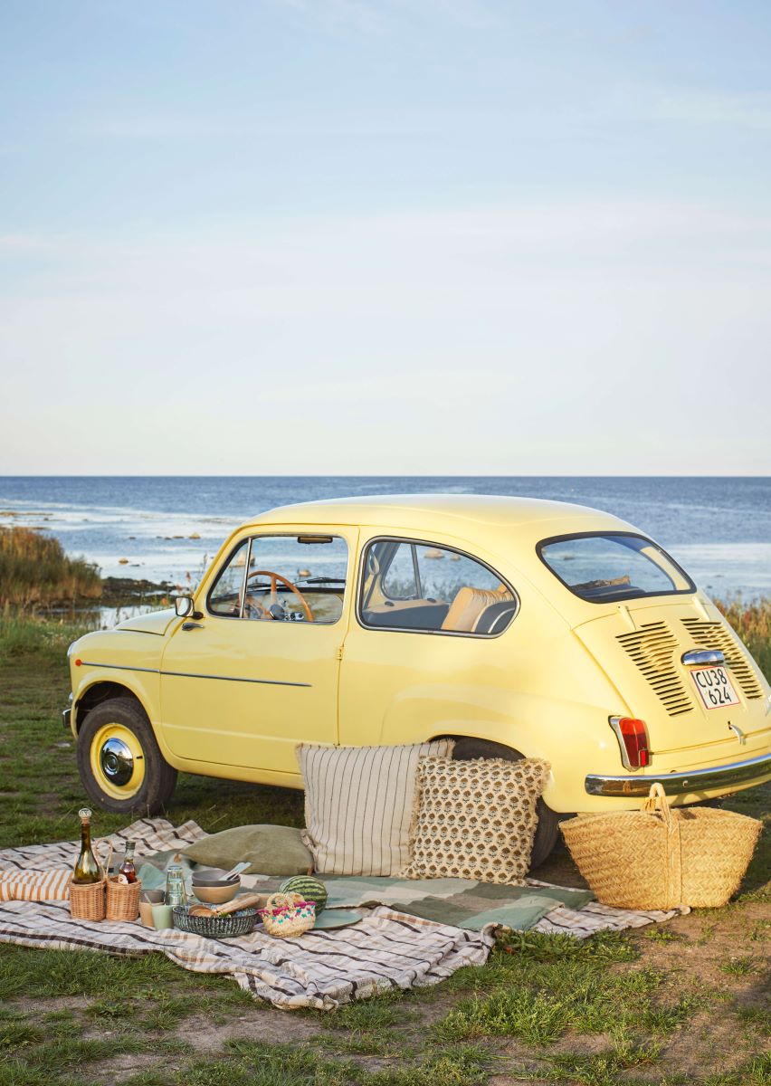 Vintage yellow car parked just behind sand dunes at the beach with a picnic set up on the grass with blankets cushions basket botte holders