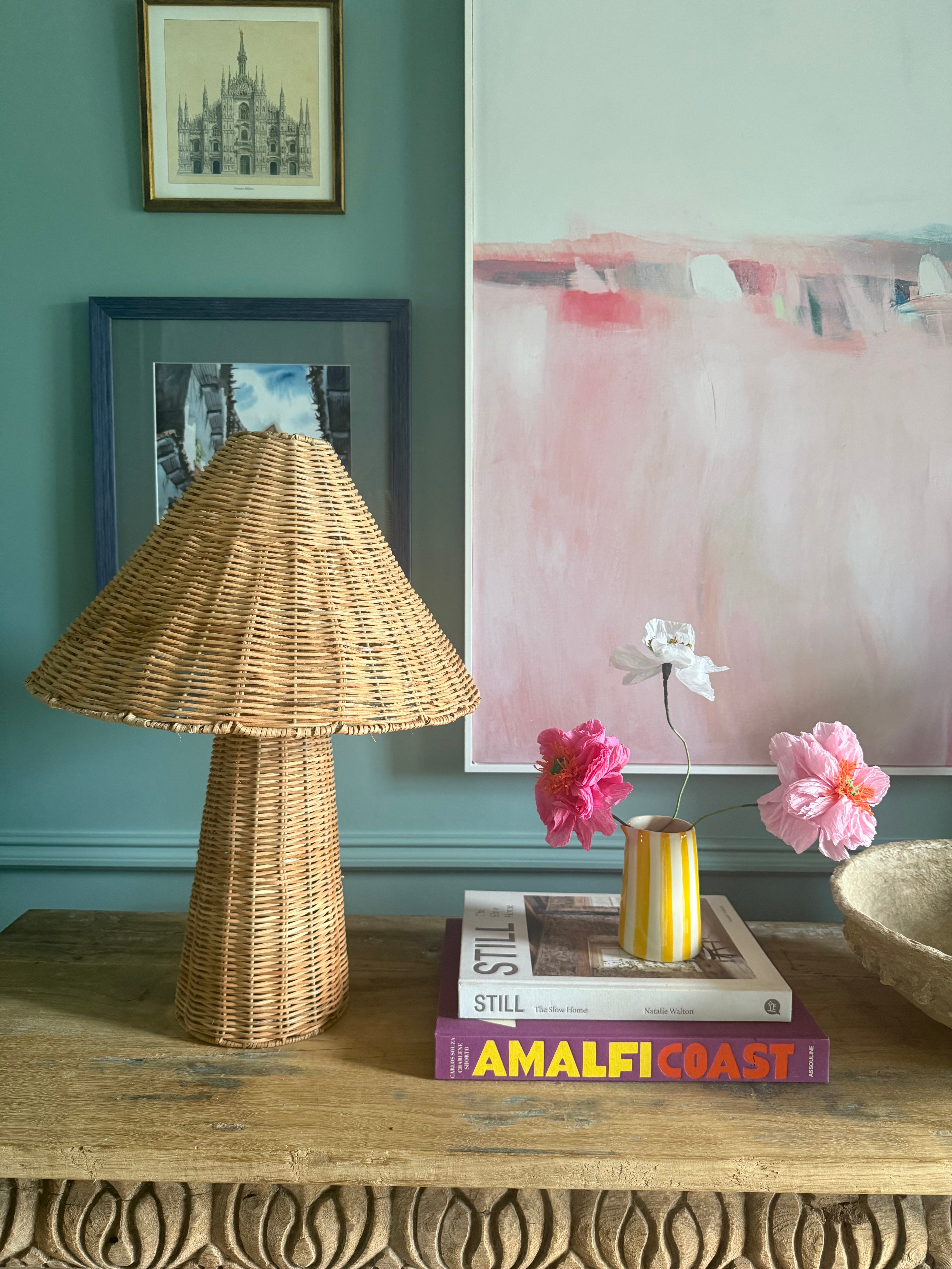 Rattan table lamp on a rustic carved console table next to stack of books with flowers and artwork on a blue-painted wall behind