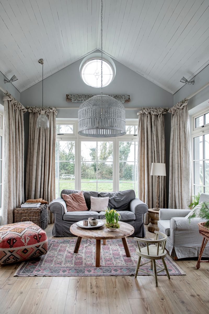 An image of a cosy living room with pitched tongue and groove ceiling, pale grey walls, large windows, linen curtians, rustic reclaimed round wood coffee table, bohemian style pouffe, gingham armchair, dark grey slip covered sofa, oversized metal factory pendant light, vintage rug, wood plank flooring