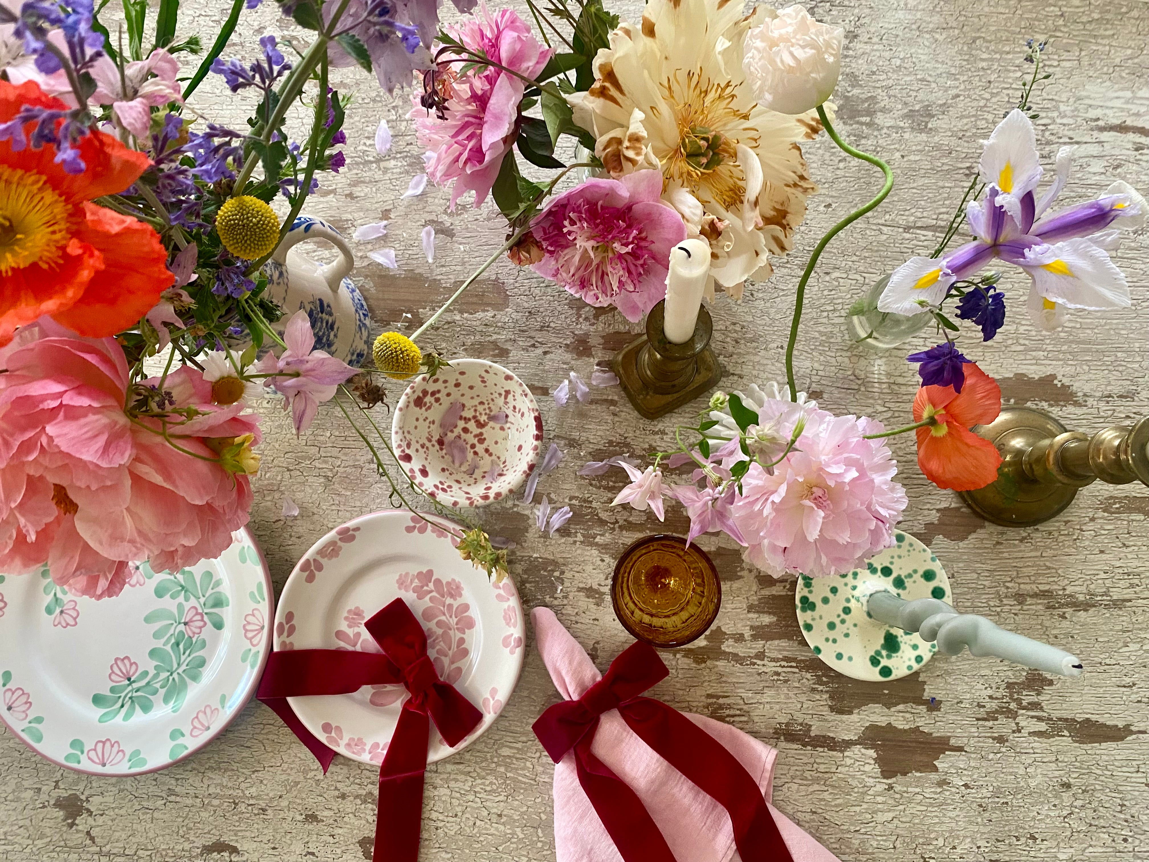 Summer table setting with bright colourful flowers in yellow, apricot, pink, orange tones with red splatter mini bowl, red velvet napkin bows, ochre glass and a dusky pink linen napkin on pink painted table
