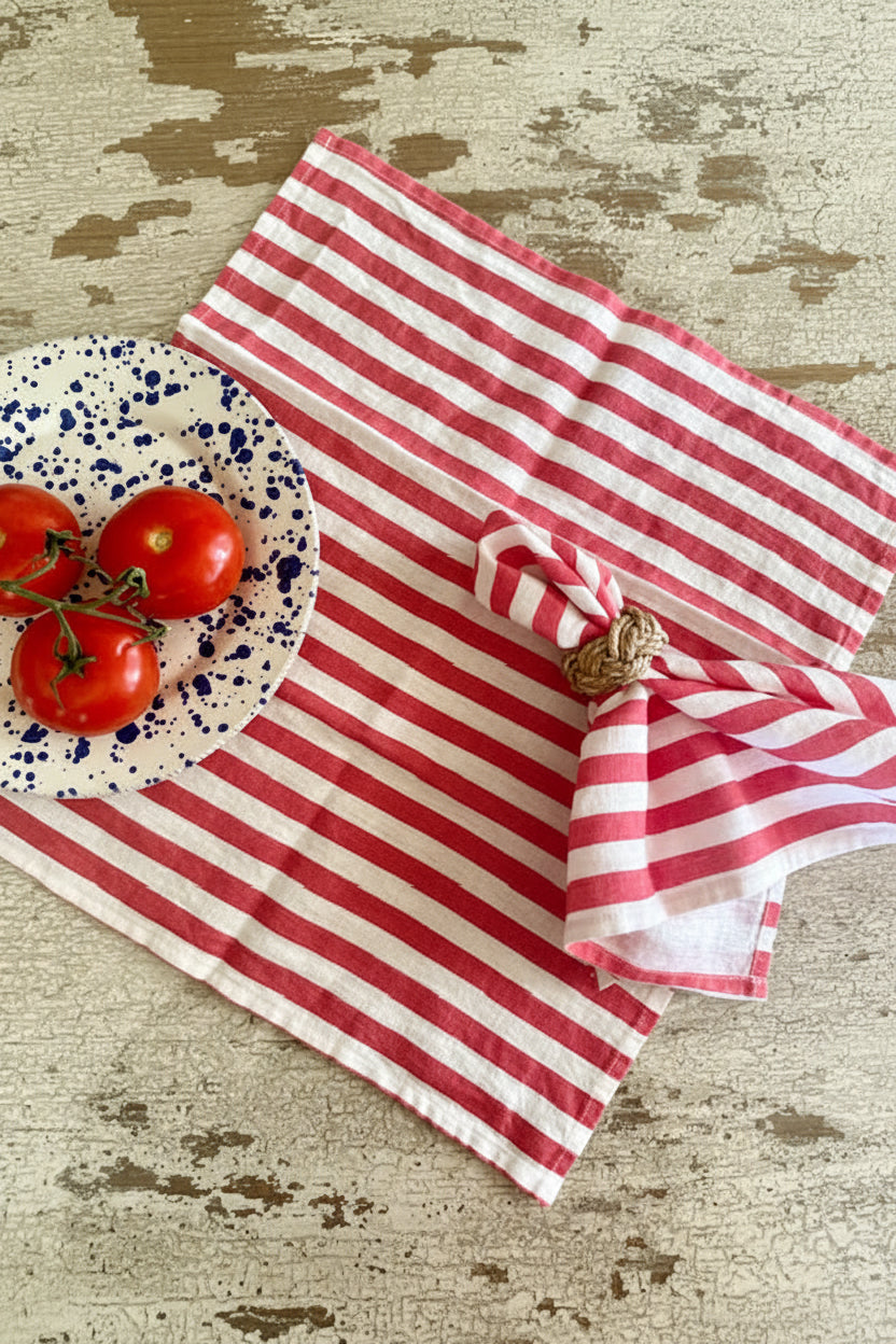 A pair of red striped linen napkins on a table with a plate of tomatoes in the background.