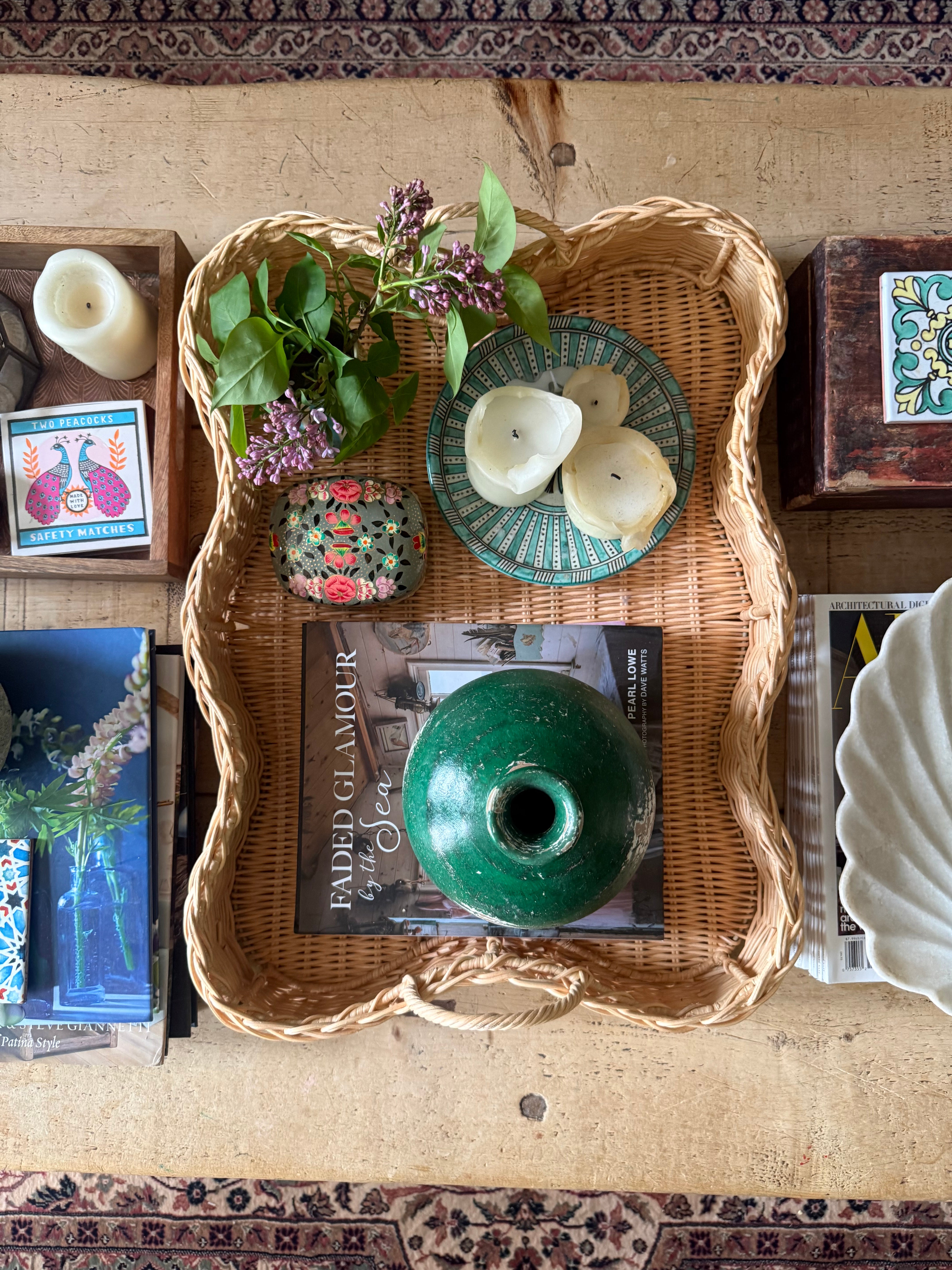 Overhead view of scalloped rattan tray on rustic coffee table with books, candles, decorative trinket box, round green vase, decorative bowls, fresh flowers