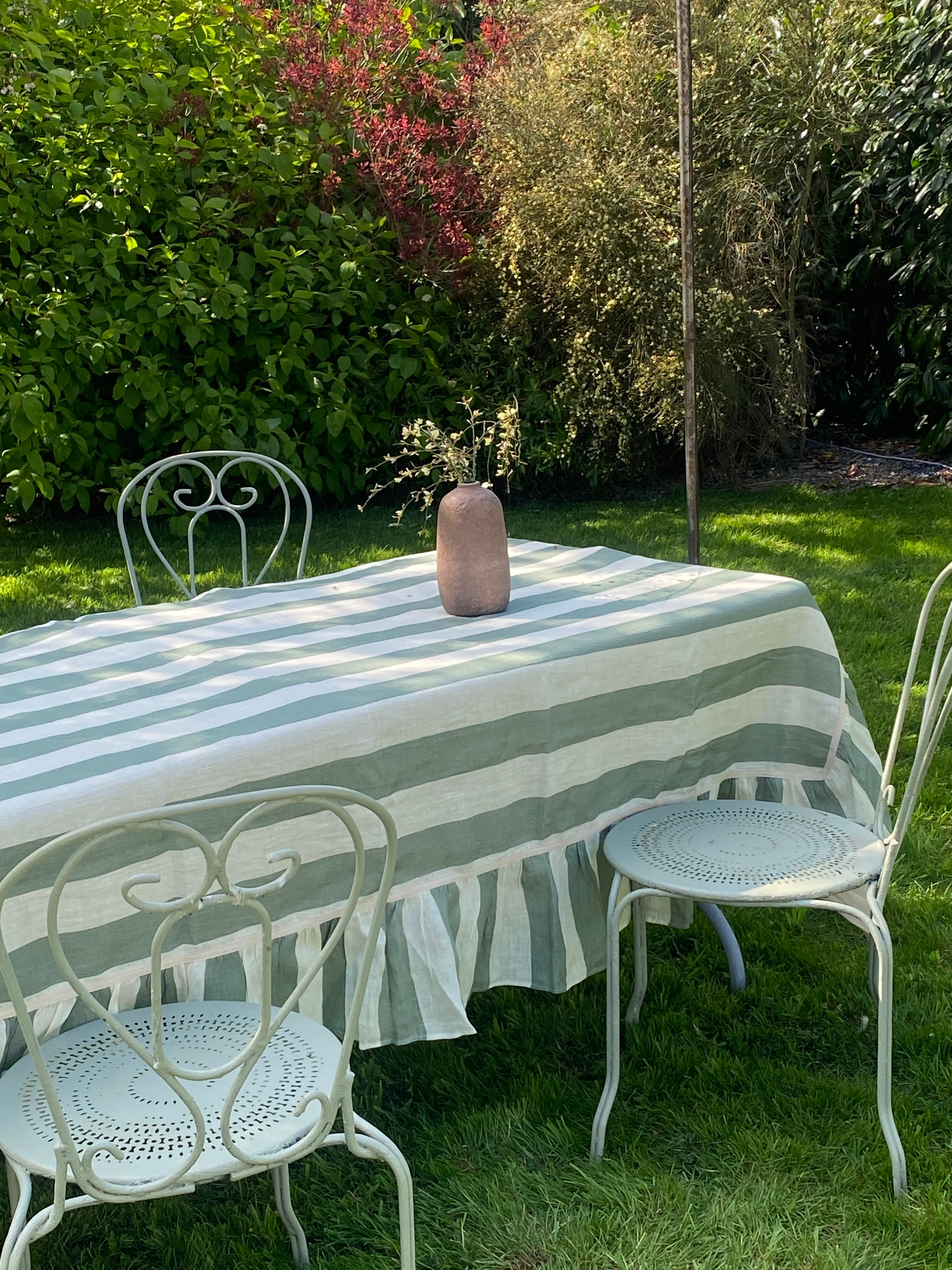 Green and white striped tablecloth with ruffled trim on a rectangular table with cream french metal scrolled chairs in a sunny garden setting