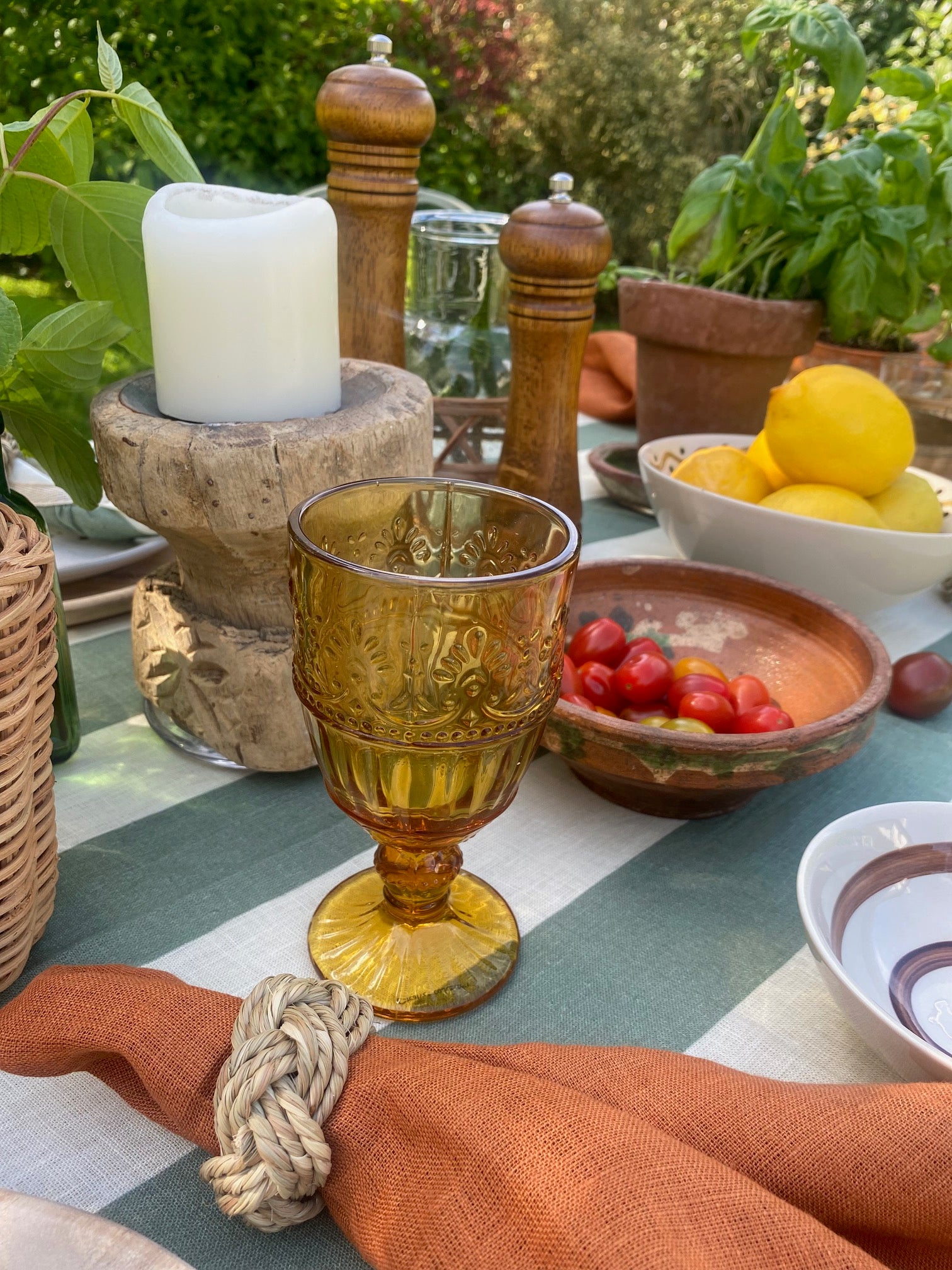 Detail of a Portofino inspired tablescape with ochre textured stemmed glass, terracotta napkin with sea grass napkin ring; vintage clay bowls, rustic wooden candle holders, potted asil and bowl of lemons on a green striped tablecloth in outdoor setting
