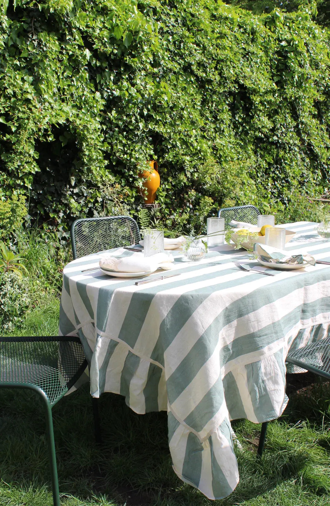 An outdoor table with sea green and white linen ruffled tablecloth against a green hedge. 