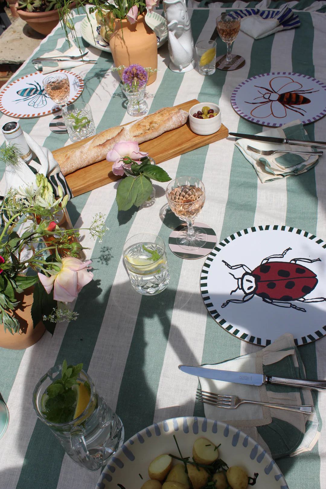 Sea green and white linen tablecloth on a sunny outdoor table setting laid with lady bug platees, bowls of food, wooden board with bread and olives, glassware and vases of natural flowers.