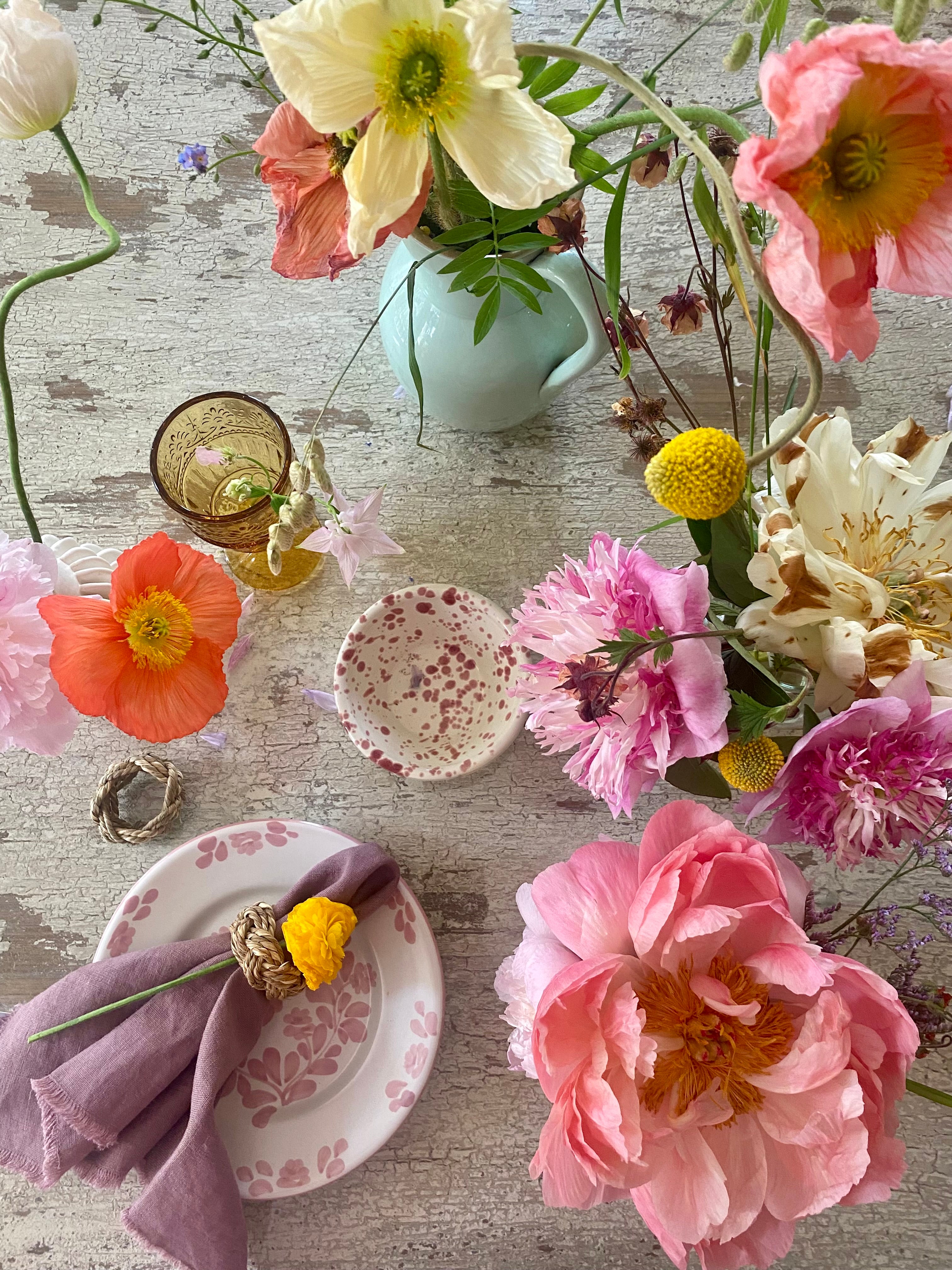 Summer table setting with bright colourful flowers in yellow, apricot, pink, orange tones with red splatter mini bowl, ochre glass and a dusky pink linen napkin on pink painted table