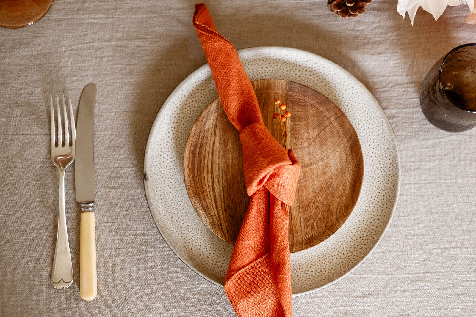 Overhead view of a place setting with terrcotta linen napkin on wood plate on textured grey brown plate on stonewashed linen tablecloth, brown glas and vintage cutlery