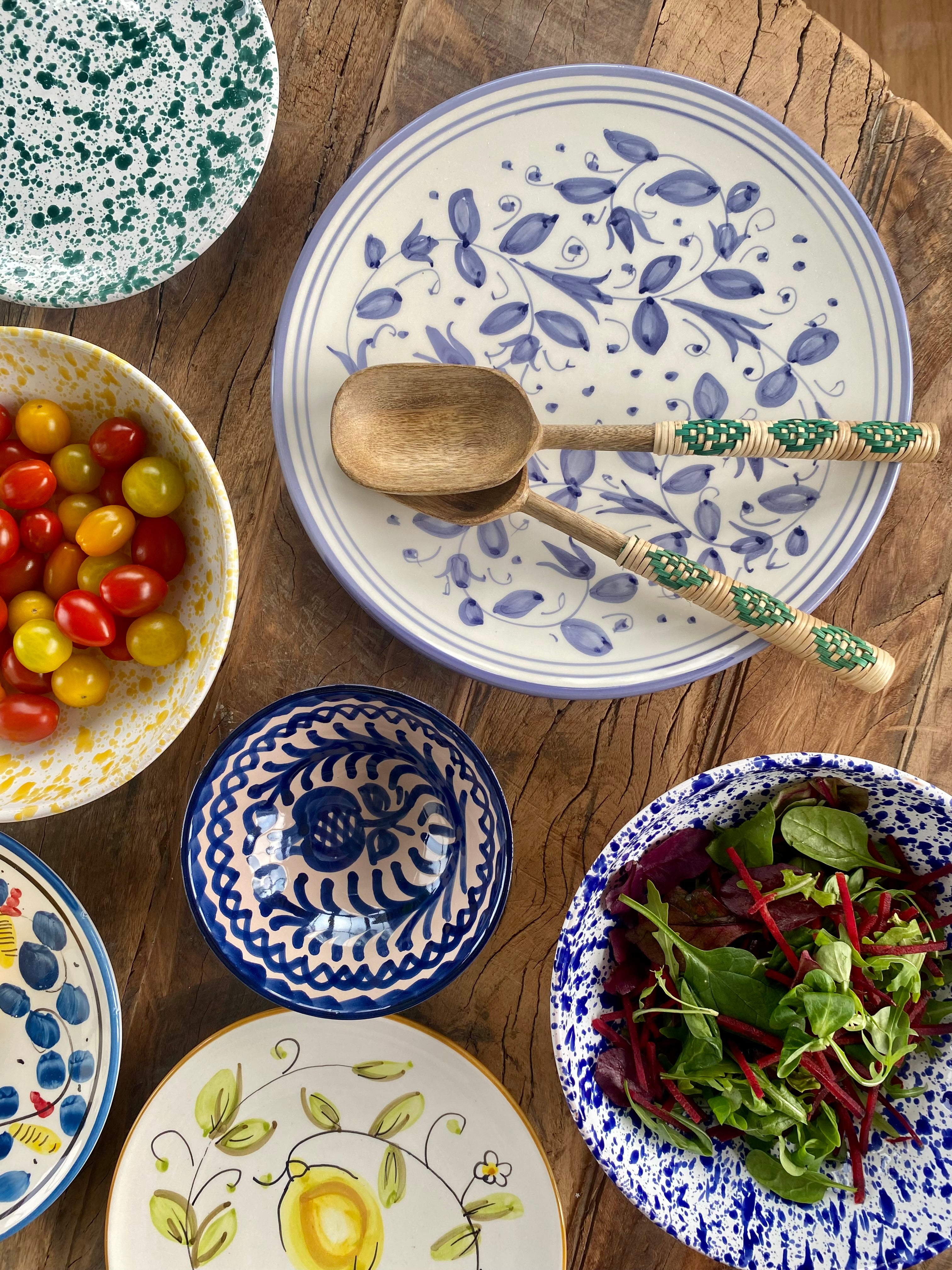 Overhead view of Spanish and Italian  plates and bowls on rustic table with salad servers, cherry tomatoes, salads. 
