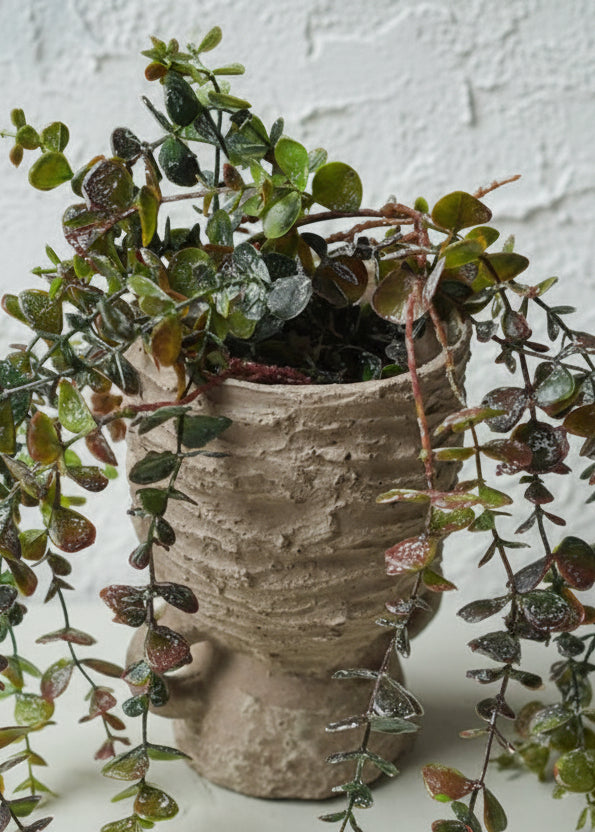 Textured raw cement vase with handles with trailing plant spilling out onto a light surface