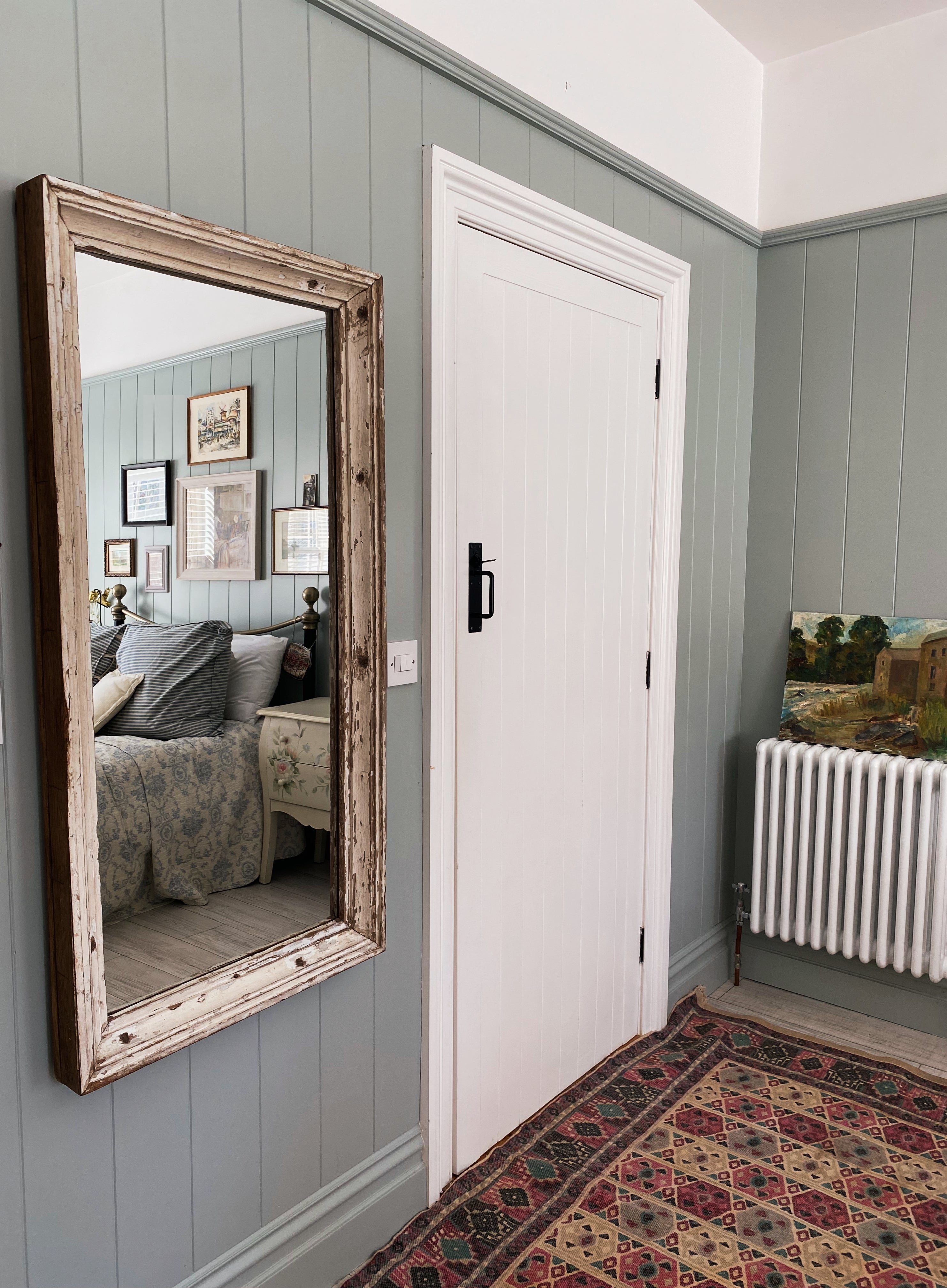 Old white reclaimed wood mirror with timeworn patina and texture hanging on a light blue painted tongue and groove panelled wall next to a white country style door with vintage rug on floor. 