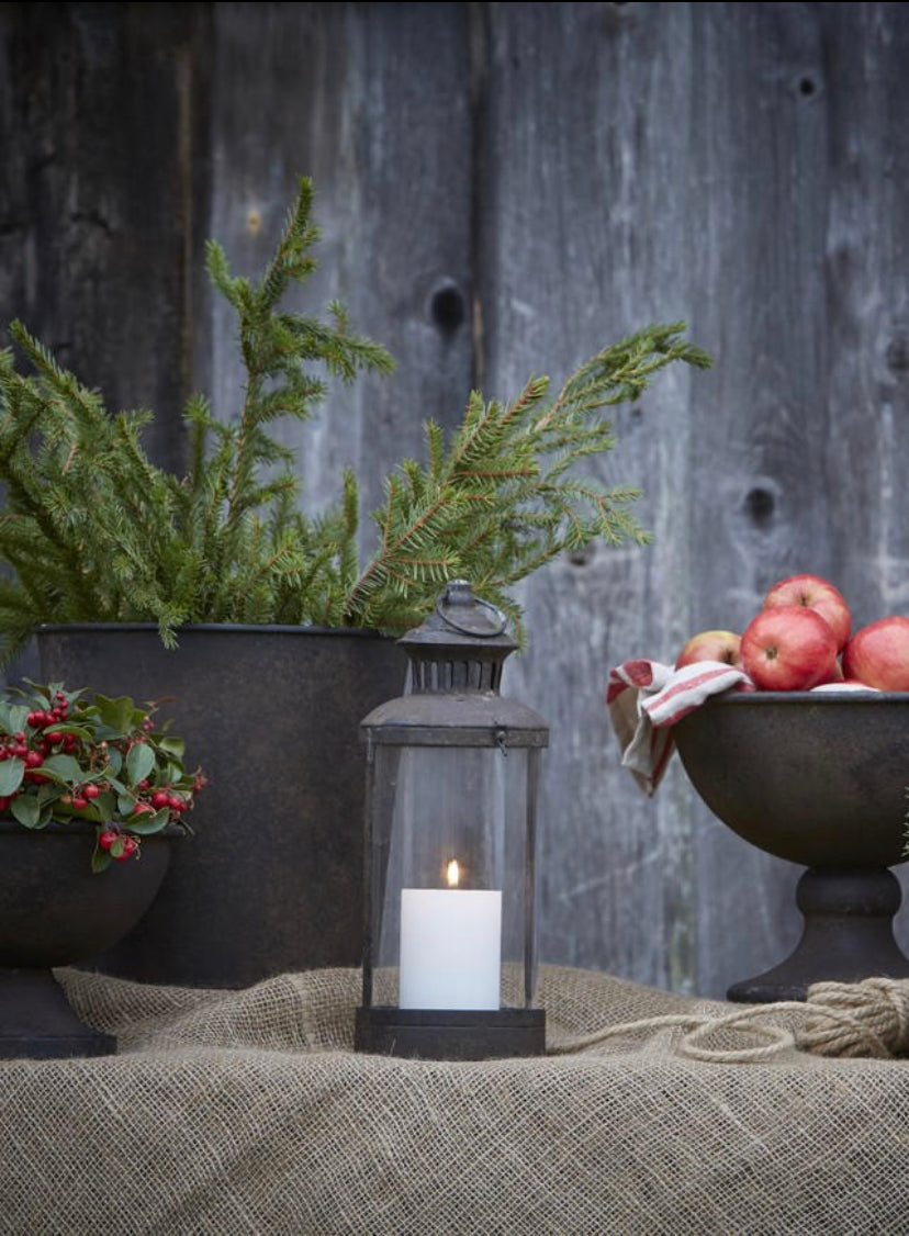 Rust lantern on table with hessian cloth and black metal footed bowls and vessels with red apples and greenery aagainst weathered grey wood fence